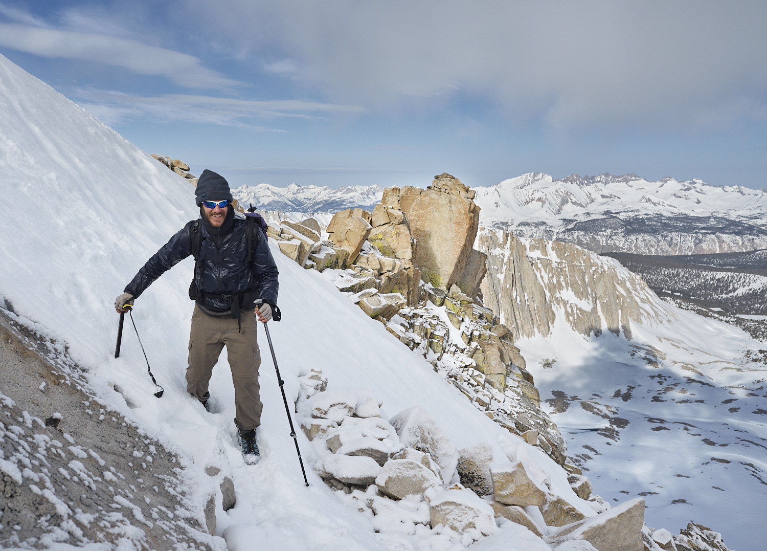 A hiker is ascending Mount Whitney. In the background is a panorama of the snow covered Sierra Nevada.