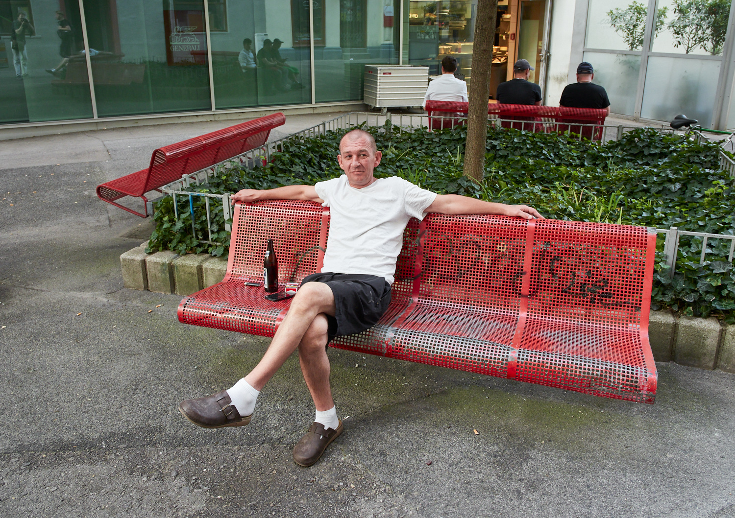 Ein Mann sitzt entspannt auf einer roten Bank mit einer Flasche Bier