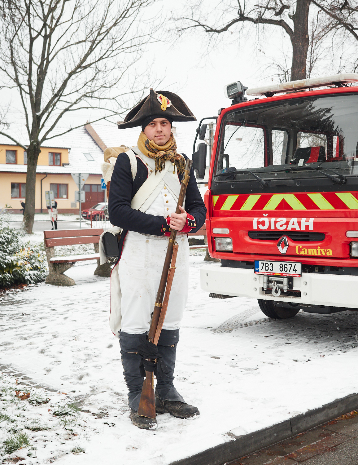 Ein junger Mann steht mit einem Gewehr in französischer Uniform aus napoleonischer Zeit vor einem Feuerwehrfahrzeug.
